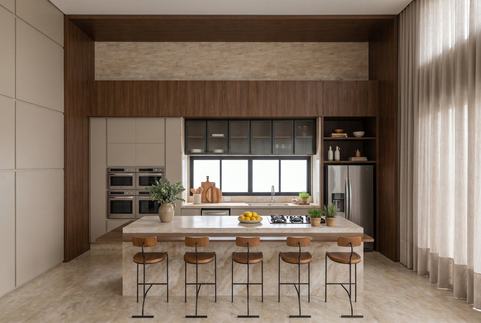 Cozinha com pórtico de madeira escura, ilha em pedra natural com cooktop e bancada de madeira, cinco banquetas, torre de fornos, aéreos de vidro canelado e cortina de linho até o teto